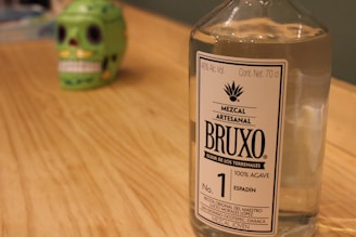 A clear glass bottle of Mezcal Artesanal with a label reading 'BRUXO Elixir de los Terrenales' is placed on a wooden surface. In the background, there is a green decorative skull with black eye sockets and white teeth, slightly out of focus.