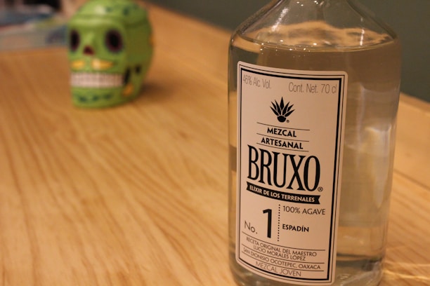 A clear glass bottle of Mezcal Artesanal with a label reading 'BRUXO Elixir de los Terrenales' is placed on a wooden surface. In the background, there is a green decorative skull with black eye sockets and white teeth, slightly out of focus.