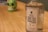 A clear glass bottle of Mezcal Artesanal with a label reading 'BRUXO Elixir de los Terrenales' is placed on a wooden surface. In the background, there is a green decorative skull with black eye sockets and white teeth, slightly out of focus.