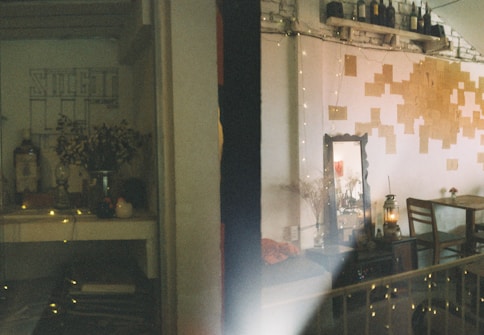 A cozy interior with soft lighting features a decorative table with a vase of flowers and small objects, such as a candle and a teapot. The wall is adorned with a grid of sticky notes or small papers. A mirror reflects part of the room, adding depth. A chair and a rustic lantern are situated near a brick wall lined with bottles.