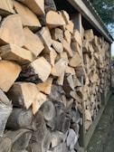 A neatly stacked pile of firewood arranged in a wooden shed. The logs are cut into uniform pieces, displaying different shades of brown and textures of bark and wood grain. The structure provides a sheltered space to keep the firewood organized and dry.