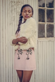 A stylish woman is standing against a vintage wooden wall with vertical lines. She is wearing a light-colored fur coat and a pink skirt with floral embroidery. Her hair is styled in a braid, and she has a confident expression while looking at the camera.
