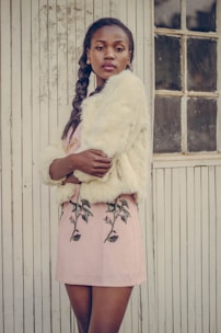 A stylish woman is standing against a vintage wooden wall with vertical lines. She is wearing a light-colored fur coat and a pink skirt with floral embroidery. Her hair is styled in a braid, and she has a confident expression while looking at the camera.