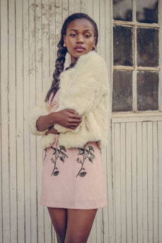 A stylish woman is standing against a vintage wooden wall with vertical lines. She is wearing a light-colored fur coat and a pink skirt with floral embroidery. Her hair is styled in a braid, and she has a confident expression while looking at the camera.