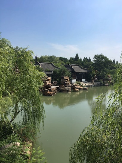A tranquil scene featuring a traditional Asian architectural structure with curved roofs, surrounded by lush greenery. The building is adjacent to a calm body of water, reflecting some of the surrounding elements. Large stones and willow trees frame the setting, suggesting a serene environment.