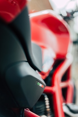 A close-up of a red motorcycle with sleek curves. The focus is on the side panel, which features a small decal with the colors of the Italian flag. Parts of the motorcycle frame and suspension are also visible.