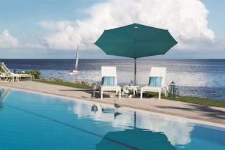 Two white lounge chairs with striped cushions are set beside a swimming pool overlooking the ocean. An aqua-colored umbrella provides shade. A sailboat is visible on the water, and a seagull stands on the pool deck. The scene is accented with decorative lanterns and lush greenery.
