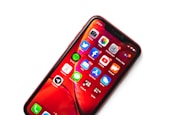 A smartphone displaying a variety of app icons against a vibrant red background. The apps include popular social media and utility apps such as Facebook, YouTube, Twitter, and Spotify. The phone screen is brightly lit, and there is a red protective case encasing the device.
