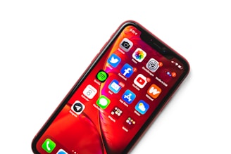 A smartphone displaying a variety of app icons against a vibrant red background. The apps include popular social media and utility apps such as Facebook, YouTube, Twitter, and Spotify. The phone screen is brightly lit, and there is a red protective case encasing the device.