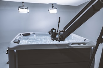 A camera mounted on a boom arm is positioned over a hot tub filled with water. The scene is illuminated by two overhead lights, creating a bright and focused setting.