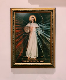 A framed religious artwork depicting a figure dressed in a white robe with a halo above the head. Radiating light in red and blue colors emanates from the heart area. The text 'JESUS I TRUST IN YOU' is written at the bottom of the image.