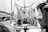 A monochrome photograph featuring a maritime setting with tightly coiled ropes hanging on the deck of a boat. The ropes are neatly arranged, and various nautical equipment and winches are visible. Surrounding the boat, other vessels and masts can be seen docked in a marina.