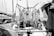 A monochrome photograph featuring a maritime setting with tightly coiled ropes hanging on the deck of a boat. The ropes are neatly arranged, and various nautical equipment and winches are visible. Surrounding the boat, other vessels and masts can be seen docked in a marina.