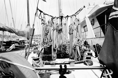 A monochrome photograph featuring a maritime setting with tightly coiled ropes hanging on the deck of a boat. The ropes are neatly arranged, and various nautical equipment and winches are visible. Surrounding the boat, other vessels and masts can be seen docked in a marina.