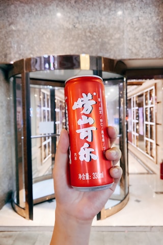 A hand is holding a red can with Chinese characters printed on it, standing in front of a modern glass revolving door.