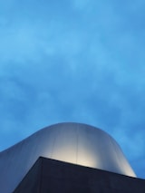 A modern architectural structure with a smooth, curved dome rises against a backdrop of a vibrant blue sky. The building facade is sleek and illuminated by soft lighting, creating a dramatic contrast with the evening sky.