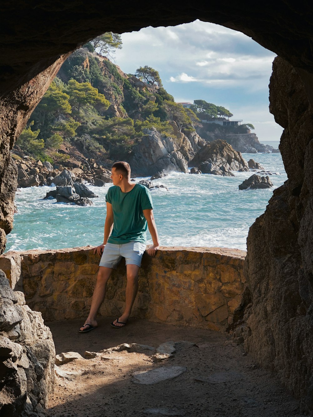man in teal t-shirt standing on rock formation near sea during daytime