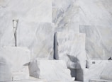 Large, rectangular slabs of white marble stacked together, showcasing natural veins and patterns in the stone surface, set against a bright and possibly harsh lighting condition.