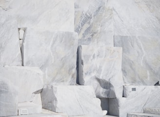 Large, rectangular slabs of white marble stacked together, showcasing natural veins and patterns in the stone surface, set against a bright and possibly harsh lighting condition.