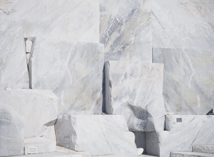 Large, rectangular slabs of white marble stacked together, showcasing natural veins and patterns in the stone surface, set against a bright and possibly harsh lighting condition.