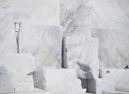 Large, rectangular slabs of white marble stacked together, showcasing natural veins and patterns in the stone surface, set against a bright and possibly harsh lighting condition.