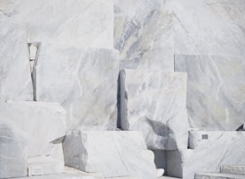 Large, rectangular slabs of white marble stacked together, showcasing natural veins and patterns in the stone surface, set against a bright and possibly harsh lighting condition.