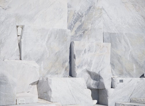 Large, rectangular slabs of white marble stacked together, showcasing natural veins and patterns in the stone surface, set against a bright and possibly harsh lighting condition.