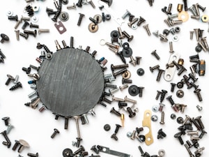 A variety of screws, nuts, and bolts are scattered on a white surface. A central round black magnet holds a number of these metallic objects, creating a circular pattern. The hardware pieces vary in color, size, and shape, appearing somewhat haphazardly arranged around the magnet.