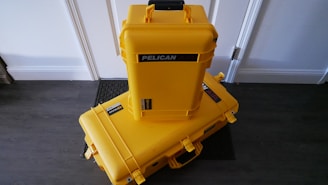 Two bright yellow protective cases labeled 'Pelican Air' are stacked on a dark wooden floor against a white wall. The larger case is lying horizontally on the floor, while the smaller case is standing vertically on top of it. Both cases have black handles and latches, with stickers indicating the brand.