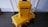 Two bright yellow protective cases labeled 'Pelican Air' are stacked on a dark wooden floor against a white wall. The larger case is lying horizontally on the floor, while the smaller case is standing vertically on top of it. Both cases have black handles and latches, with stickers indicating the brand.