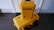 Two bright yellow protective cases labeled 'Pelican Air' are stacked on a dark wooden floor against a white wall. The larger case is lying horizontally on the floor, while the smaller case is standing vertically on top of it. Both cases have black handles and latches, with stickers indicating the brand.