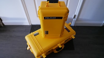 Two bright yellow protective cases labeled 'Pelican Air' are stacked on a dark wooden floor against a white wall. The larger case is lying horizontally on the floor, while the smaller case is standing vertically on top of it. Both cases have black handles and latches, with stickers indicating the brand.