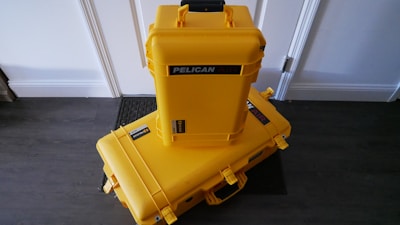 Two bright yellow protective cases labeled 'Pelican Air' are stacked on a dark wooden floor against a white wall. The larger case is lying horizontally on the floor, while the smaller case is standing vertically on top of it. Both cases have black handles and latches, with stickers indicating the brand.