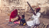 A scene featuring a man in traditional attire playing a stringed musical instrument, seated on the ground. He wears a colorful turban and is accompanied by a small child who looks relaxed and a woman with her face covered by a red veil, sitting on a patterned fabric. The setting appears rustic, with a textured stone wall in the background.