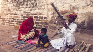 A scene featuring a man in traditional attire playing a stringed musical instrument, seated on the ground. He wears a colorful turban and is accompanied by a small child who looks relaxed and a woman with her face covered by a red veil, sitting on a patterned fabric. The setting appears rustic, with a textured stone wall in the background.