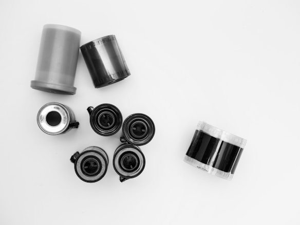 A collection of film canisters and film rolls arranged on a plain white background. The image includes several black and grey cylindrical canisters and rolls of film, some with visible film strips.