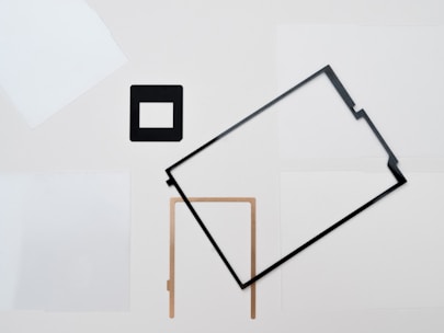 A minimalist composition features various geometric shapes arranged on a neutral background. These include a black square frame and a rectangular frame, along with a L-shaped object, all spread irregularly across the image.