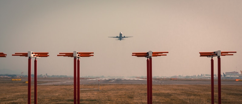 An airplane is taking off or landing on a runway, surrounded by tall red lights. The sky is overcast, creating a muted atmosphere.
