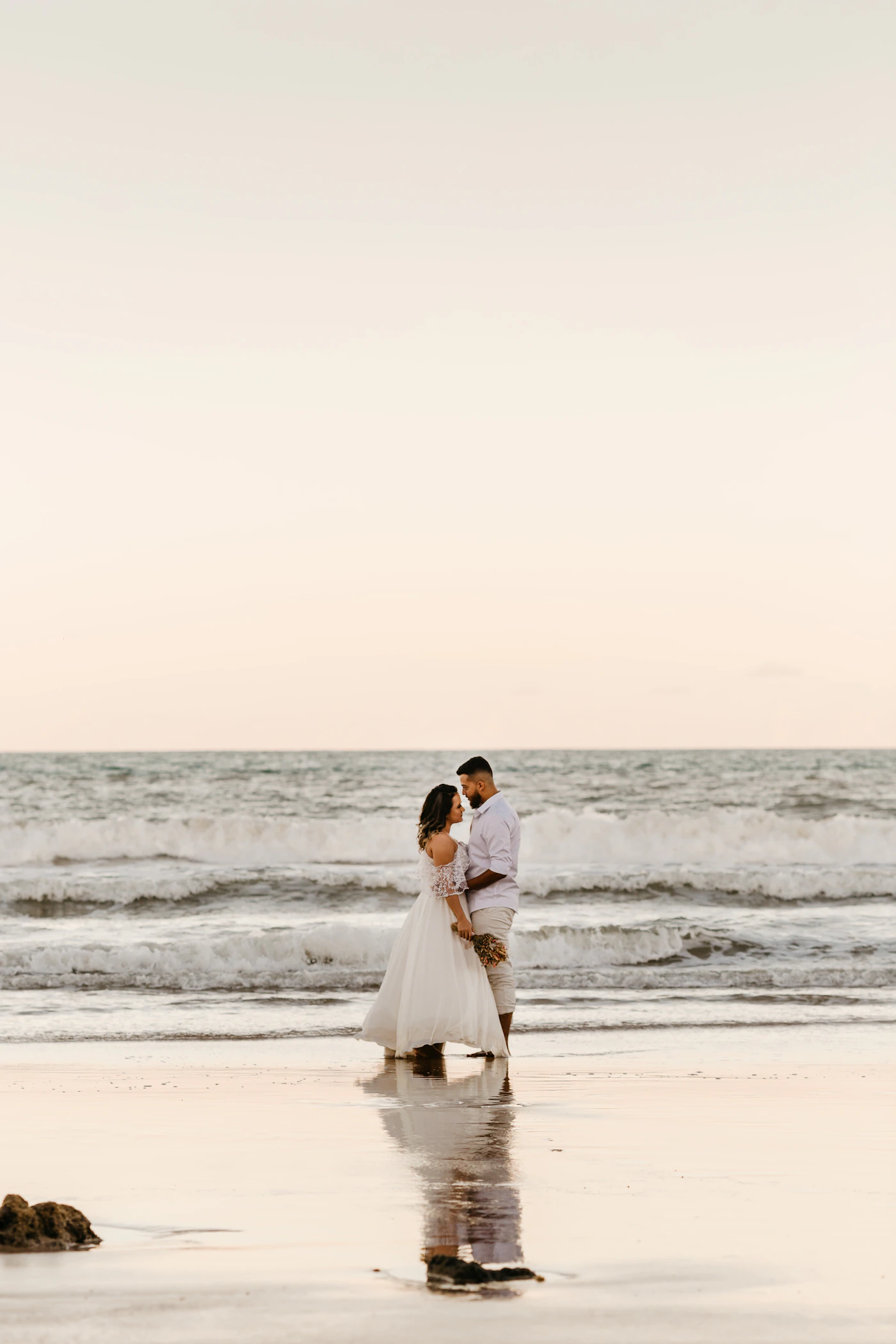 Couple in elopement attire standing at the shoreline of a Colombian Pacific beach with waves coming in around their feet and the jungle behind them