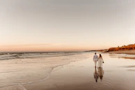 A candid moment of a couple walking hand in hand along a sandy beach at dusk.