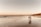 A couple walks hand in hand along a sandy beach during sunset, with gentle waves lapping at their feet. The sky is a gradient of warm colors and the reflection of the couple is visible on the wet sand. Cliffs line the distant shoreline, adding depth to the scene.