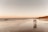 A couple walks hand in hand along a sandy beach during sunset, with gentle waves lapping at their feet. The sky is a gradient of warm colors and the reflection of the couple is visible on the wet sand. Cliffs line the distant shoreline, adding depth to the scene.
