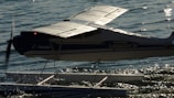 A small seaplane with a prominent wing structure is positioned on calm water. The plane's body is marked with the words 'GRAND TUNDRA' and it features pontoons for landing on water. The propeller is slightly blurred, indicating motion. The sunlight reflects off the water's surface, creating a shimmering effect.