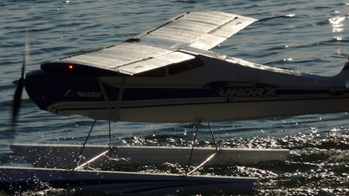 A small seaplane with a prominent wing structure is positioned on calm water. The plane's body is marked with the words 'GRAND TUNDRA' and it features pontoons for landing on water. The propeller is slightly blurred, indicating motion. The sunlight reflects off the water's surface, creating a shimmering effect.