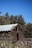 A rustic wooden shed with a metal roof is set against a backdrop of dense green trees under a clear blue sky. The structure shows signs of age with weathered wooden panels and surrounded by tall grasses and wildflowers.