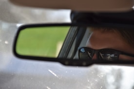 A close-up of a car's rearview mirror reflects a person wearing dark sunglasses, focusing on driving. The mirror captures part of the face and the area outside the car, while the rest of the image is blurred.
