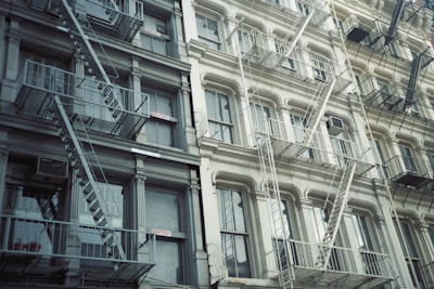 A series of old-style apartment buildings with multiple fire escape ladders attached to them. The buildings feature large windows, and the structure is made up of gray and off-white materials. Some windows have air conditioning units installed. The buildings have decorative columns and molding details.