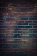 A wall composed of dark teal rectangular bricks with reddish-brown mortar lines creating a grid-like pattern. The surface of the bricks reflects some light, giving them a slightly glossy appearance, which contributes to variations in the shade and texture.