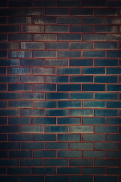 A wall composed of dark teal rectangular bricks with reddish-brown mortar lines creating a grid-like pattern. The surface of the bricks reflects some light, giving them a slightly glossy appearance, which contributes to variations in the shade and texture.