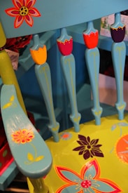 A vividly painted wooden chair featuring floral patterns. The chair has a turquoise color with red, yellow, and dark purple accent flowers. Detailed floral designs are present on both the seat and the armrest, adding a playful touch.